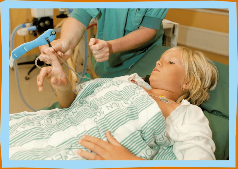 Blond pojke ligger i en säng. Sjuksköterska sätter en liten blå lampa på pojkens fingertopp.