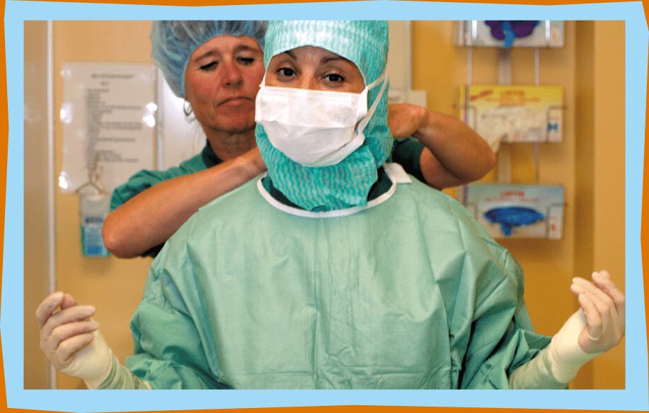 Kirurg i operationskläder, latexhandskar, operationsmössa och munskydd står framför en sjuksköterska som hjälper till med påklädning.
