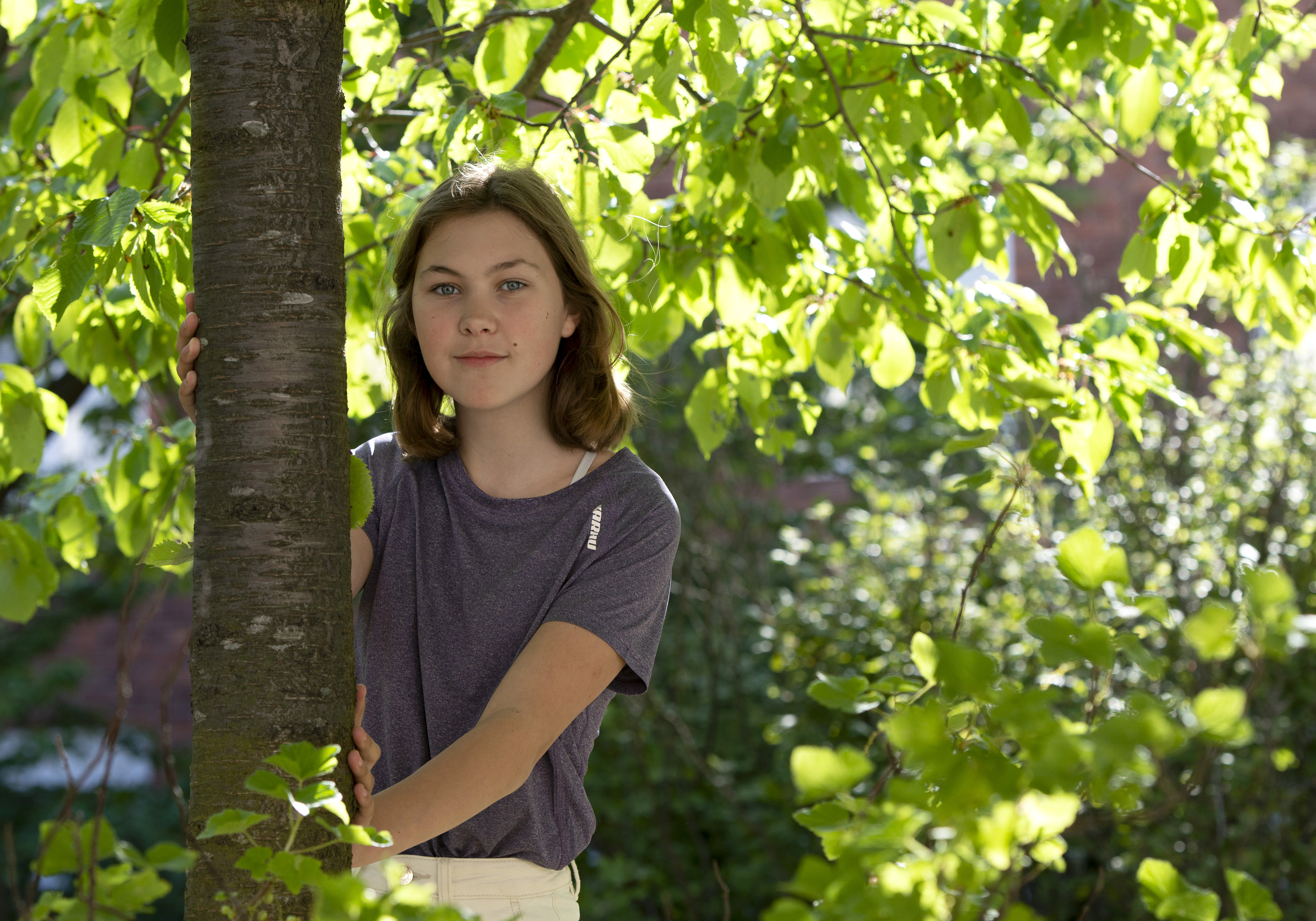 Mathilda står lutad mot ett träd, med gröna grenar och solljus i bakgrunden.