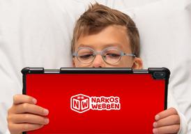 En pojke med runda blå glasögon ligger i sin säng och kollar på en röd Ipad.