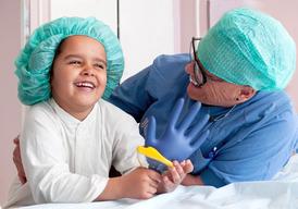 En flicka och en sjuksköterska i gröna operationsmössor sitter bredvid varann och skrattar. Flickan har en blå handske i handen.