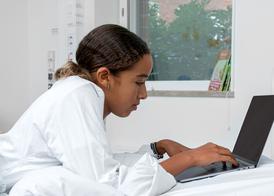 En tonårstjej ligger på mage i sin sjukhussäng och kollar på en dator.