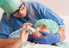En flicka ligger på ett operationsbord med en andningsmask över ansiktet. Hon tittar på en sjuksköterska som sitter bakom henne.