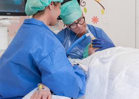 En patient ligger på ett operationsbord och andas i en syremask. Patientens mamma sitter bredvid opperationsbordet.