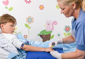 En pojke sitter i en säng. En sjuksköterska sätter in en plastslang i ett blodkärl på pojkens vänstra hand.