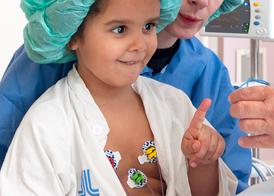 En flicka sitter i sin mammas knä i ett operationsrum. Hon sträcker ut sitt pekfinger mot en syremätare och ser nyfiken ut.