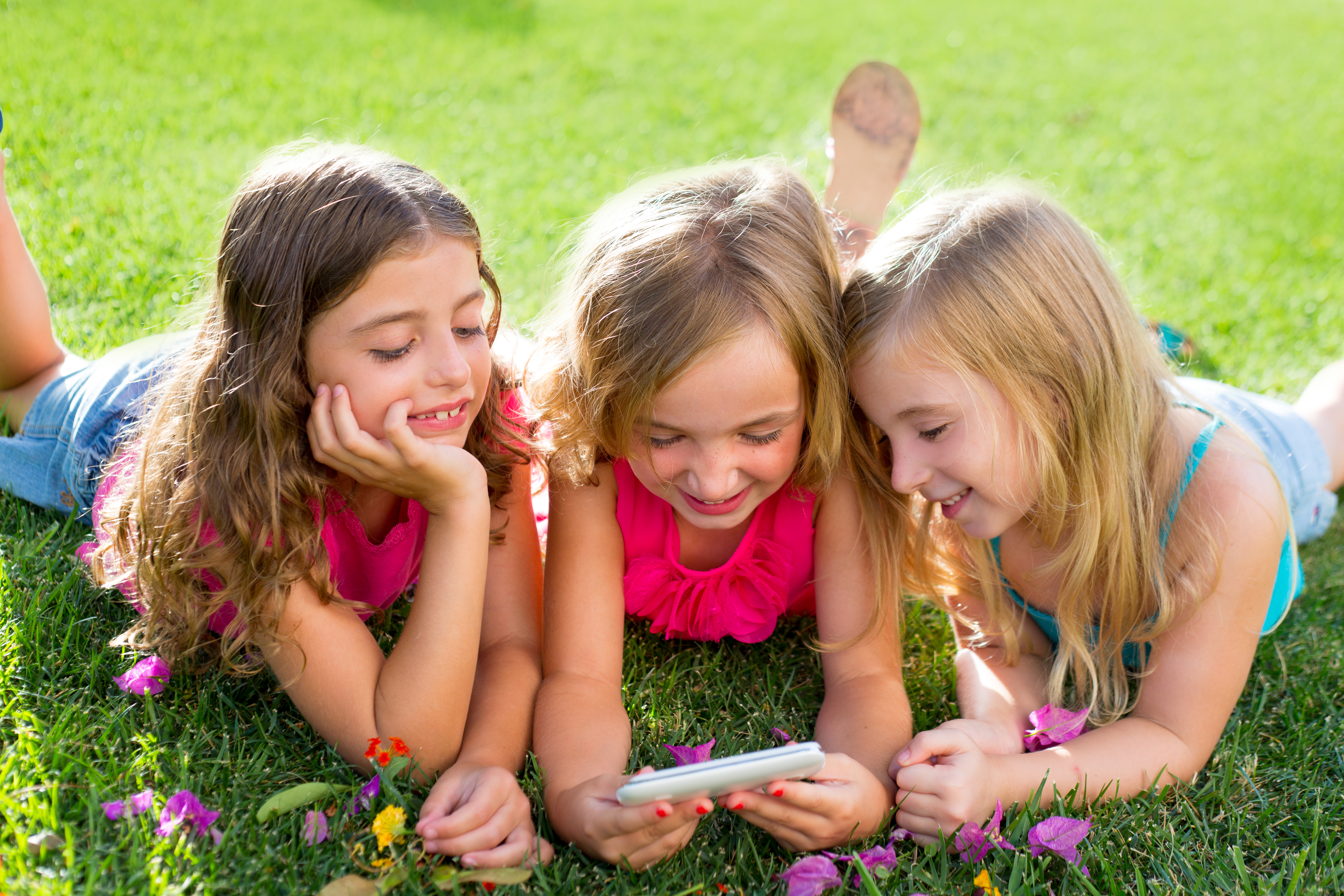 Tre flickor ligger i gräset och tittar på en mobiltelefon. 