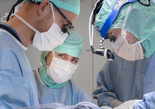 Tre kirurger opererar. En kvinnlig kirurg tittar in i kameran.
